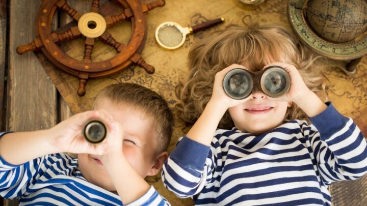 Hygge Children - Two children laying on their backs with binoculars on top of an old map and globe nearby