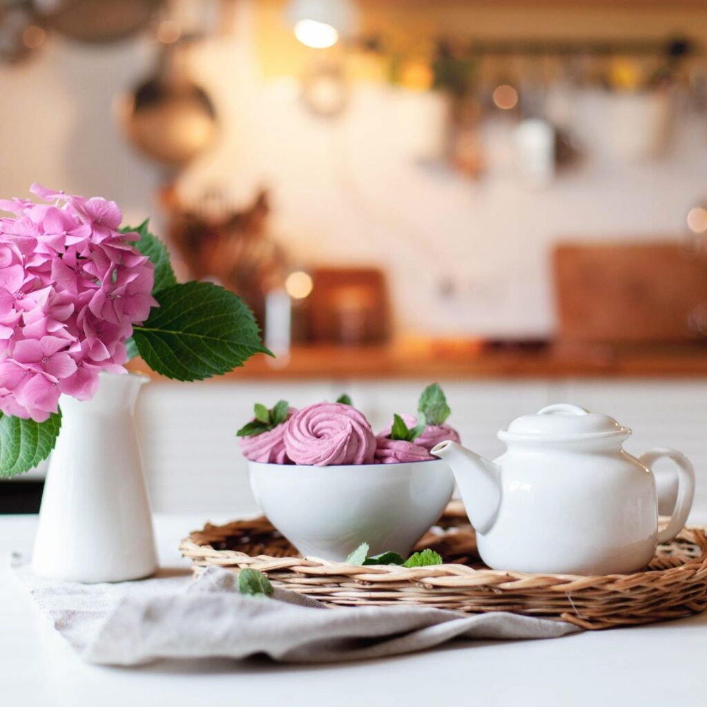 21 Secrets for a Tidy Home - Wicker Basket tray with a white teapot and a white bowl of sweets and a white vase with pink hydrangea. Background the kitchen.