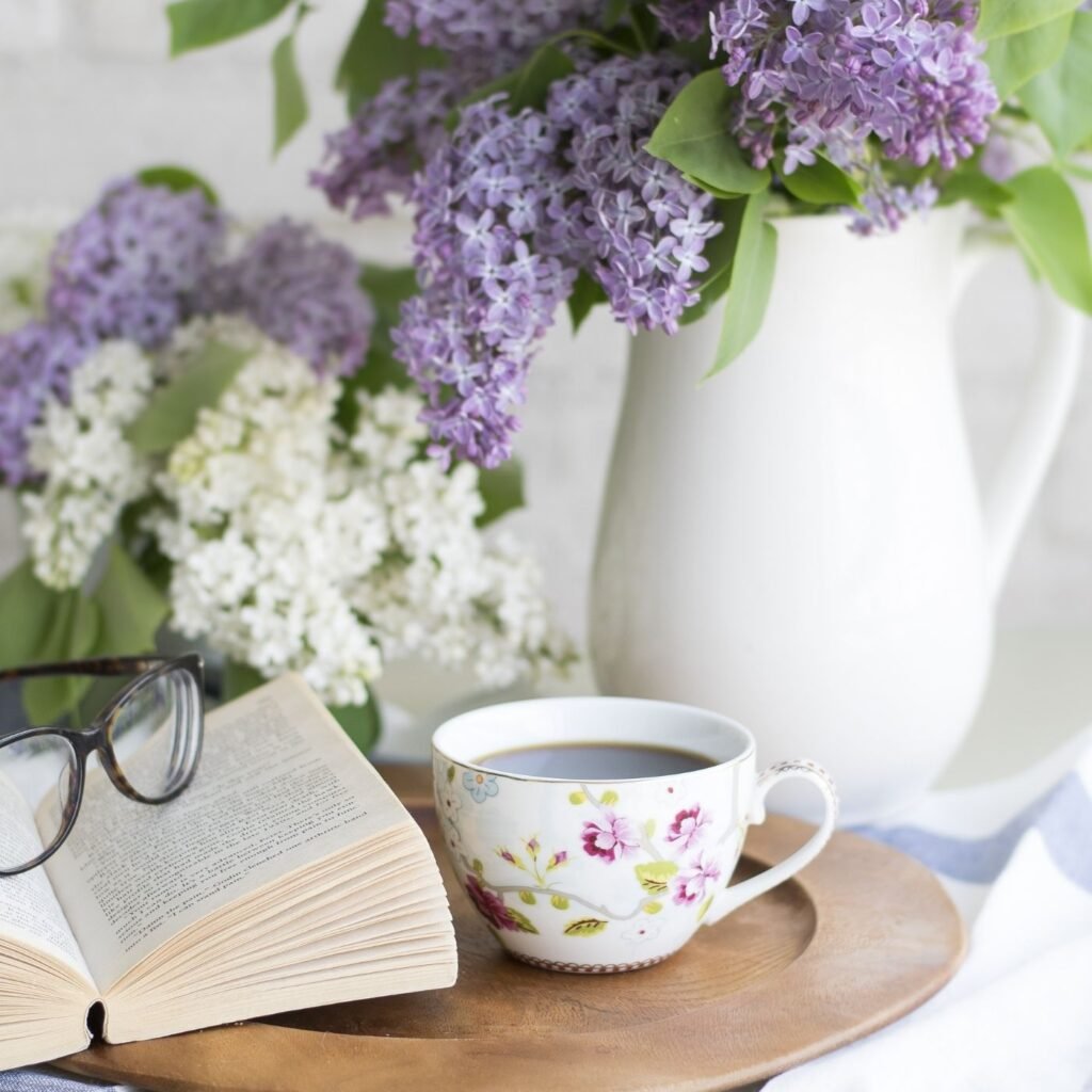 Wooden tray with mug of coffee, book, glasses, and pitcher with lilacs.